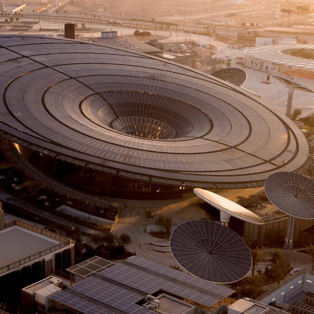 L'image montre un bâtiment architectural moderne et futuriste, avec un design en forme d'anneau ou de soucoupe volante. Le toit est en grande partie vitré, permettant à la lumière naturelle d'entrer. Des structures circulaires, semblables à des panneaux solaires ou des œuvres d'art, entourent le bâtiment, ajoutant à son aspect innovant. Le paysage environnant semble désertique, avec des infrastructures modernes visibles à l'arrière-plan, indiquant un environnement urbain en développement. La lumière douce du coucher du soleil donne une ambiance chaleureuse à la scène.