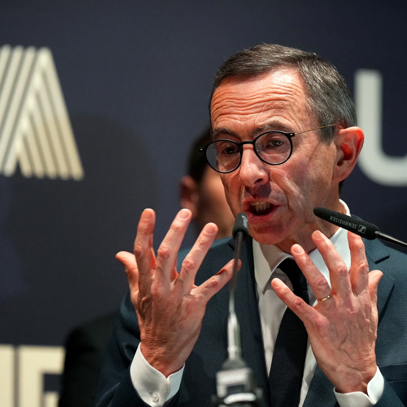 الصورة تظهر شخصًا يرتدي نظارات ويبدو أنه يتحدث بحماس، حيث يستخدم يديه للتعبير عن أفكاره. خلفه توجد خلفية داكنة تحمل شعار يبدو أنه مرتبط بمؤسسة أو منظمة. الأضواء تركز عليه مما يعطي انطباعًا بأهمية الحدث الذي يتحدث فيه.