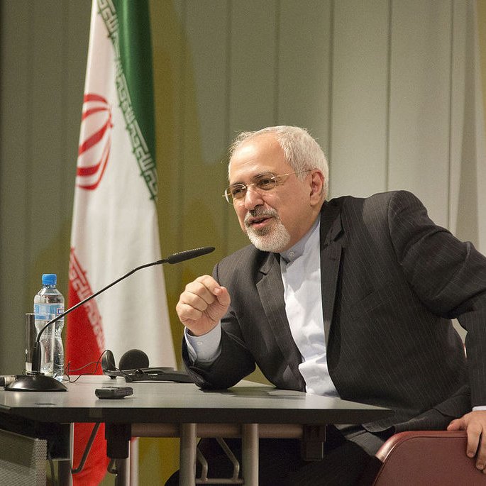 L'image montre un homme assis à une table de conférence, parlant avec animation. Il est habillé en costume, avec une chemise claire. À l'arrière-plan, on peut voir des drapeaux, y compris celui de l'Iran et celui des États-Unis. L'homme semble s'adresser à un public, probablement en répondant à des questions ou en faisant une déclaration importante.