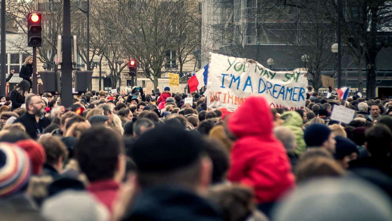 L'image montre une grande foule rassemblée, probablement pour une manifestation ou une marche. On peut voir des personnes de tous âges, certaines portant des écharpes ou des bonnets, tandis que d'autres sont vêtues de manteaux. Au centre, il y a une pancarte sur laquelle est écrit : "YOU MAY SAY I'M A DREAMER BUT I'M NOT THE ONLY ONE". Des drapeaux français sont visibles, ce qui suggère un contexte patriotique ou une revendication sociale. L'atmosphère semble être à la fois solide et engagée.