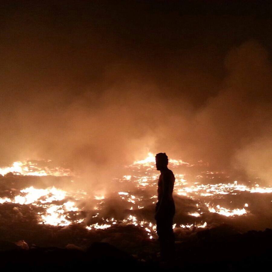 L'image montre un paysage de feu et de fumée. On voit un homme de silhouette, se tenant debout au premier plan, face à un vaste champ d'incendie. Le sol est enflammé, et une épaisse fumée s'élève, créant une atmosphère sombre et chaotique. Les flammes et la lumière orangée contrastent avec l'obscurité environnante, soulignant la gravité de la situation.