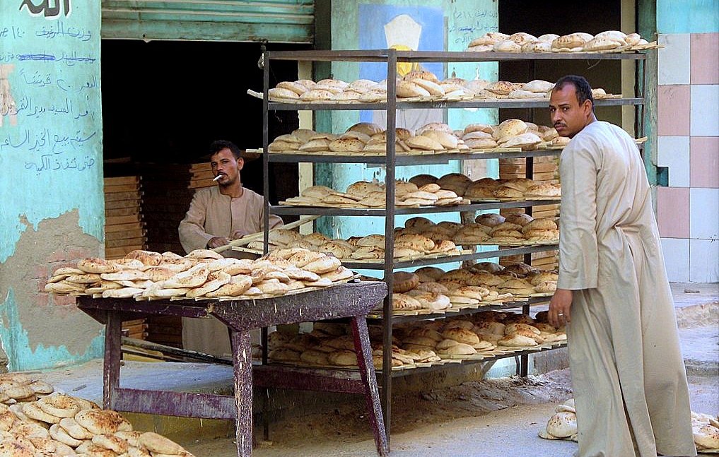 L'image montre une scène de la vie quotidienne dans une boulangerie. Il y a deux hommes : l'un est debout derrière un chariot portant des pains, tandis que l'autre semble en train d'observer ou d'interagir avec des pains empilés sur des étagères. Les pains sont de forme ronde et semblent frais, sortant tout juste du four. En arrière-plan, on aperçoit un mur avec des écritures et des graffiti, donnant une atmosphère typique d'un marché local. Le paysage autour est urbain, avec des bâtiments qui semblent usés par le temps.