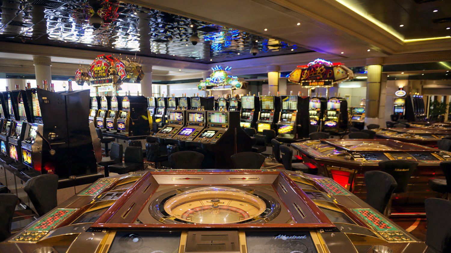 تصویر یک کازینو را نشان می‌دهد که در آن میزهای قمار و دستگاه‌های اسلات قرار دارند. در وسط تصویر، میز رولت با چرخ و دکمه‌های مختلف دیده می‌شود. در پس‌زمینه، دستگاه‌های اسلات و دیگر میزهای قمار به صورت منظم چیده شده‌اند. نورپردازی کازینو جذاب و رنگارنگ است و فضای عمومی، نوعی شلوغی و هیجان را به وجود آورده است.