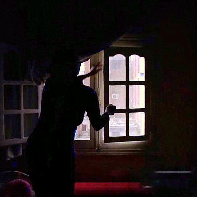 L'image montre une silhouette se tenant près d'une fenêtre dans une pièce sombre. La lumière entre par la fenêtre, illuminant partiellement l'espace environnant, tandis que des détails tels que les murs et les meubles sont à peine visibles dans l'ombre. La silhouette semble en mouvement, ajoutant une ambiance dynamique et mystérieuse à la scène. Les rideaux sont légèrement ouverts, laissant entrevoir l'extérieur.