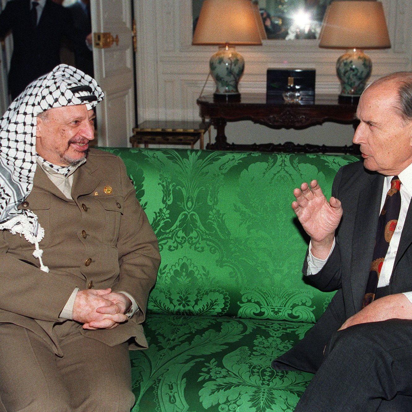 L'image montre deux hommes assis sur un canapé vert décoré. L'un des hommes porte un costume beige avec un keffieh, tandis que l'autre est en costume sombre avec une cravate à motifs. Ils semblent engagés dans une conversation animée. En arrière-plan, on aperçoit des éléments de mobilier et une lumière douce, suggérant un cadre intérieur élégant.