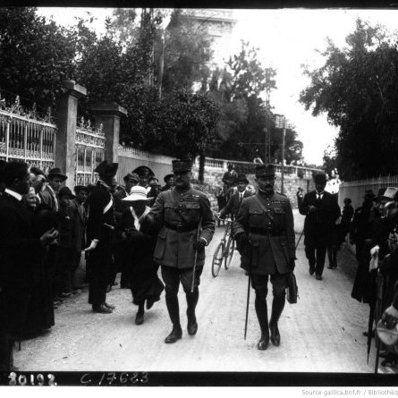 L'image montre une rue entourée de personnes, dont des militaires en uniforme. Deux officiers marchent au premier plan, semblant discuter. On aperçoit des civils sur les côtés, certains regardant vers les officiers. L'ambiance semble solennelle, et l'environnement est arboré, avec des bâtiments en arrière-plan. Les vêtements des personnes indiquent une époque historique. L'image a une qualité noire et blanche, typique des photographies anciennes.