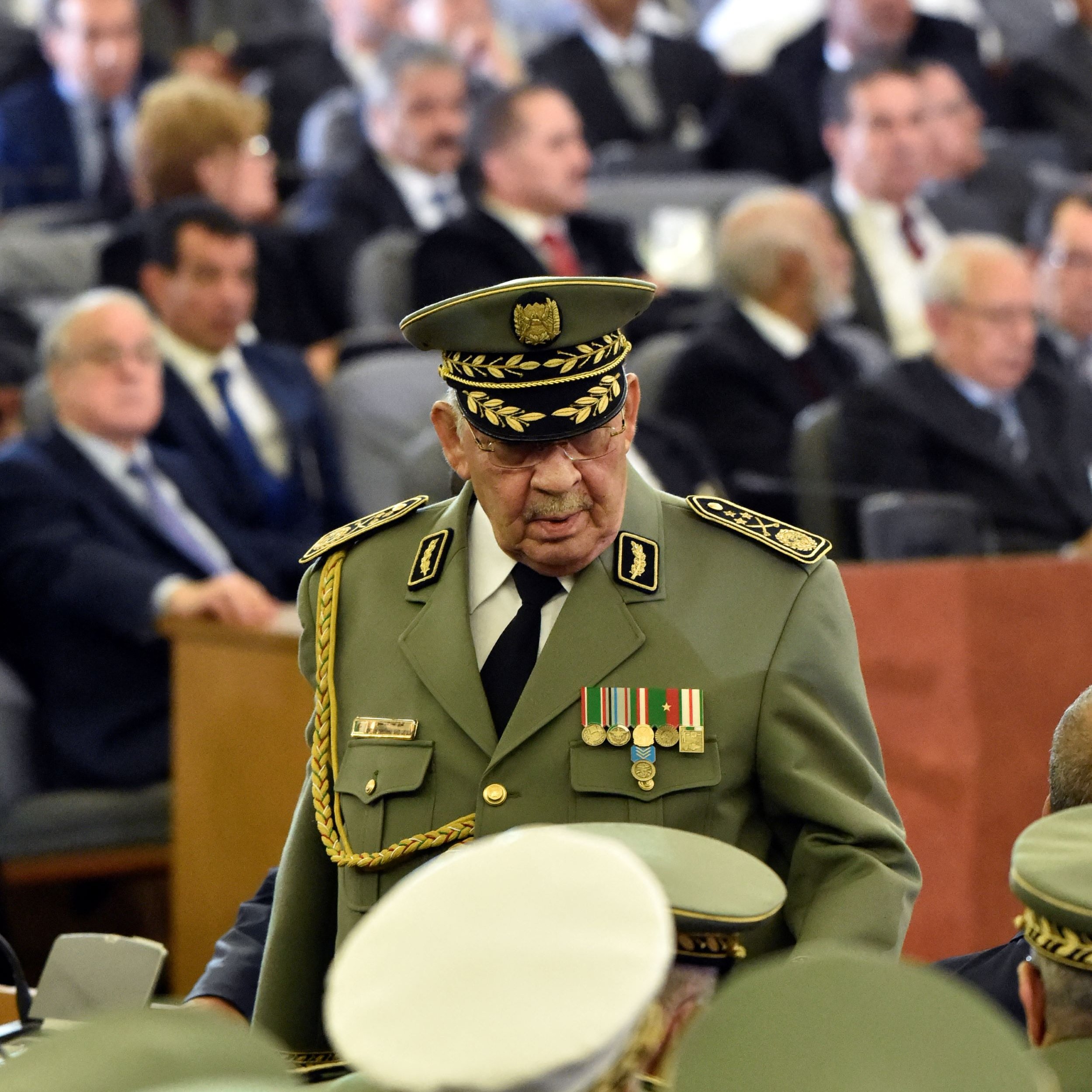 L'image montre un groupe de personnes réunies dans une grande salle, avec des sièges disposés en rangées. Au premier plan, un homme en uniforme militaire avec un chapeau prononcé, orné de médailles, se déplace parmi la foule. Dans l'arrière-plan, on aperçoit d'autres hommes et femmes, certains en costume, d'autres portant des turbans, tous attentifs à l'événement. L'atmosphère semble formelle, suggérant une occasion importante ou une cérémonie.