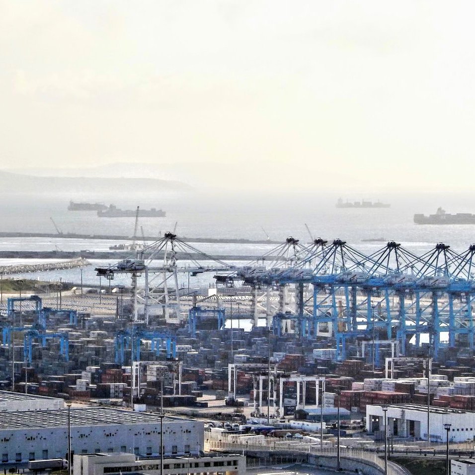 L'image montre un port maritime avec des conteneurs empilés, des grues de chargement et plusieurs navires au large. On peut voir des bâtiments et des installations portuaires. L'horizon est légèrement brumeux, créant une atmosphère calme, et des bateaux de différentes tailles sont visibles sur la mer. L'ensemble dépeint une scène de transport maritime et de logistique.