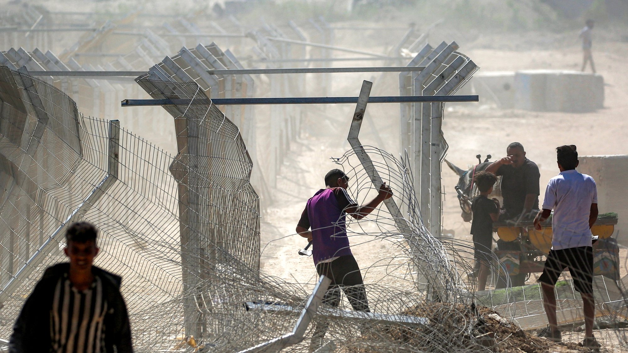 تصویر نشان‌دهنده مردانی است که در حال عبور از حصاری فلزی و برقراری یا تخریب موانع هستند. گرد و غبار در هواست.