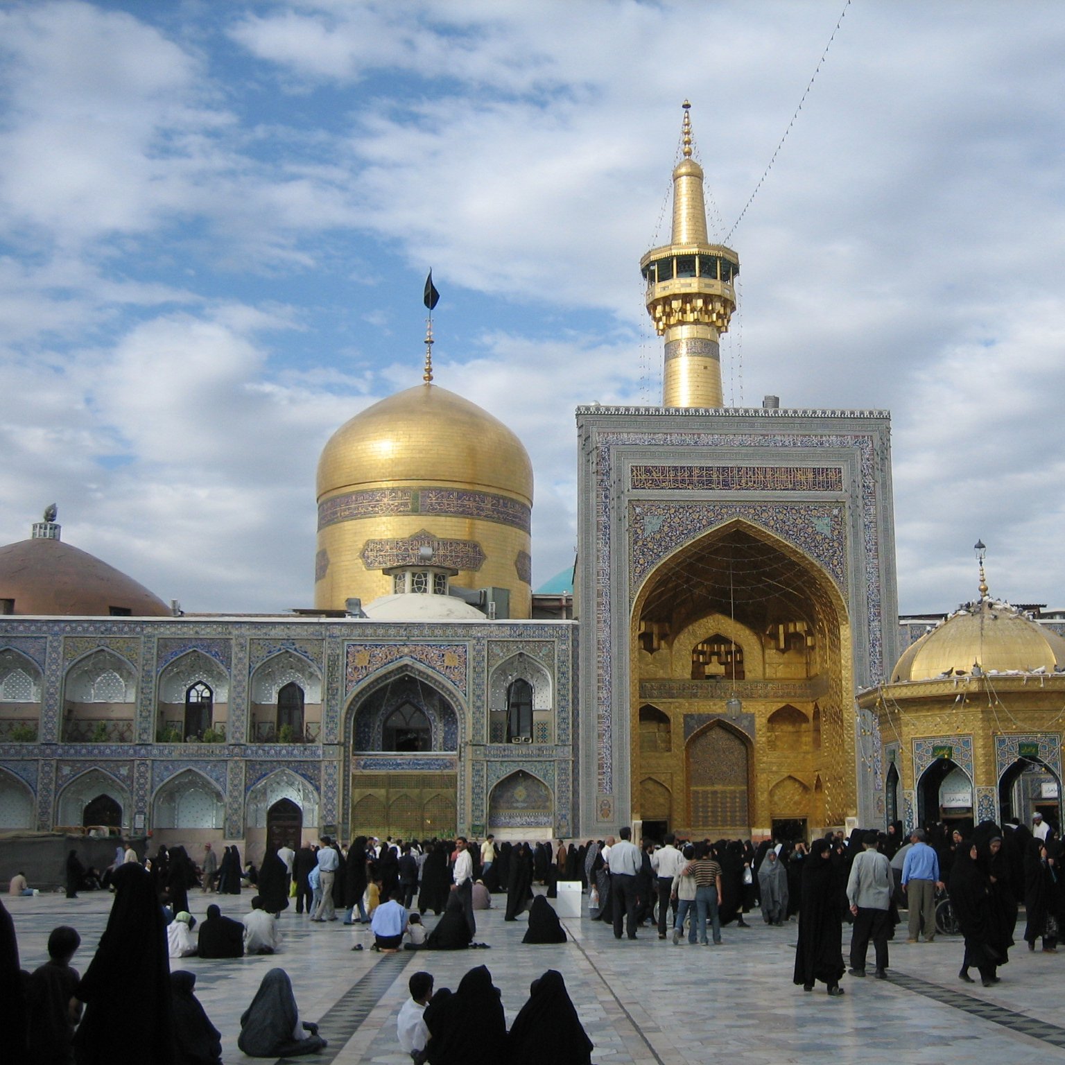 الصورة تظهر ضريحًا ذو قبة ذهبية وجنبة زاهية، حيث يحيط به عدد من الأشخاص الذين يرتدون ملابس تقليدية طويلة. السماء في الخلفية ملبدة بالغيوم، مما يعطي جوًا روحانيًا. المعمار مزخرف بشكل جميل بالزخارف الإسلامية، ويبدو أن المكان يعج بالزوار.