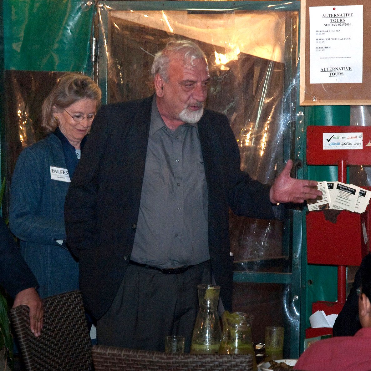 L'image montre un groupe de personnes rassemblées dans un espace qui semble être un restaurant ou une salle de réunion. Un homme au centre, avec des cheveux gris et portant une chemise noire, semble être en train de parler ou de gesticuler. À sa gauche, une femme avec des cheveux ondulés est debout et regardent vers lui. Un autre homme, assis, écoute attentivement, tandis que d'autres personnes sont également présentes à la table. L'environnement est décontracté, avec des éléments typiques de la restauration, tels que des verres et des boissons sur la table.