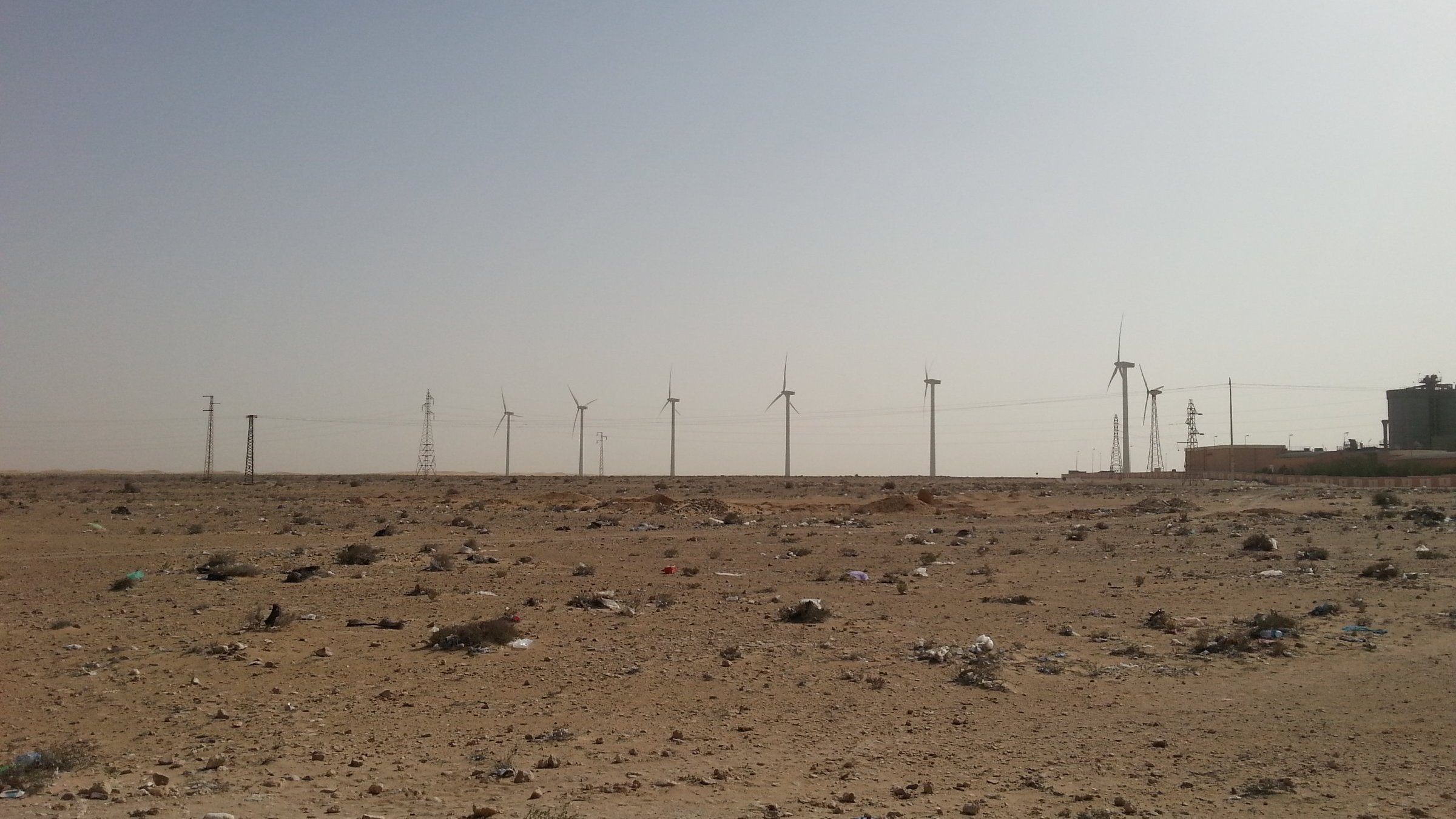 L'image montre un paysage désertique, caractérisé par un terrain aride avec des débris, probablement des déchets, éparpillés sur le sol. À l'arrière-plan, on peut voir plusieurs éoliennes qui s'élèvent vers le ciel clair. Des pylônes électriques sont également visibles, ajoutant une touche industrielle à ce paysage naturel. Le ciel est dégagé, ce qui accentue l'effet désertique de la scène.