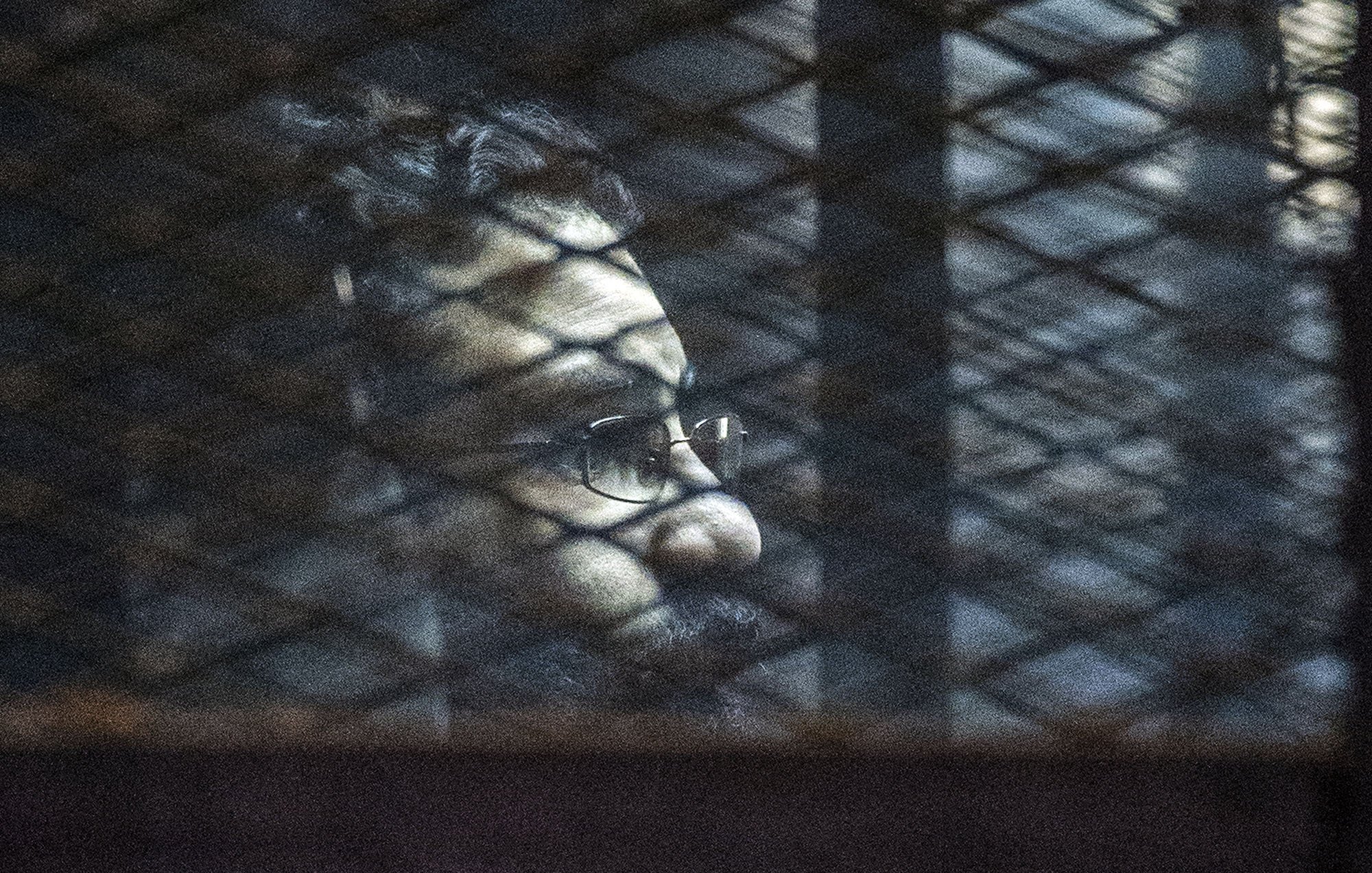 L'immagine mostra un uomo visto attraverso una rete metallica. La luce crea ombre sul suo viso, evidenziando la sua espressione seria. Indossa occhiali e ha una barba, mentre lo sfondo risulta scuro e indistinto, contribuendo a un'atmosfera di introspezione o solitudine.