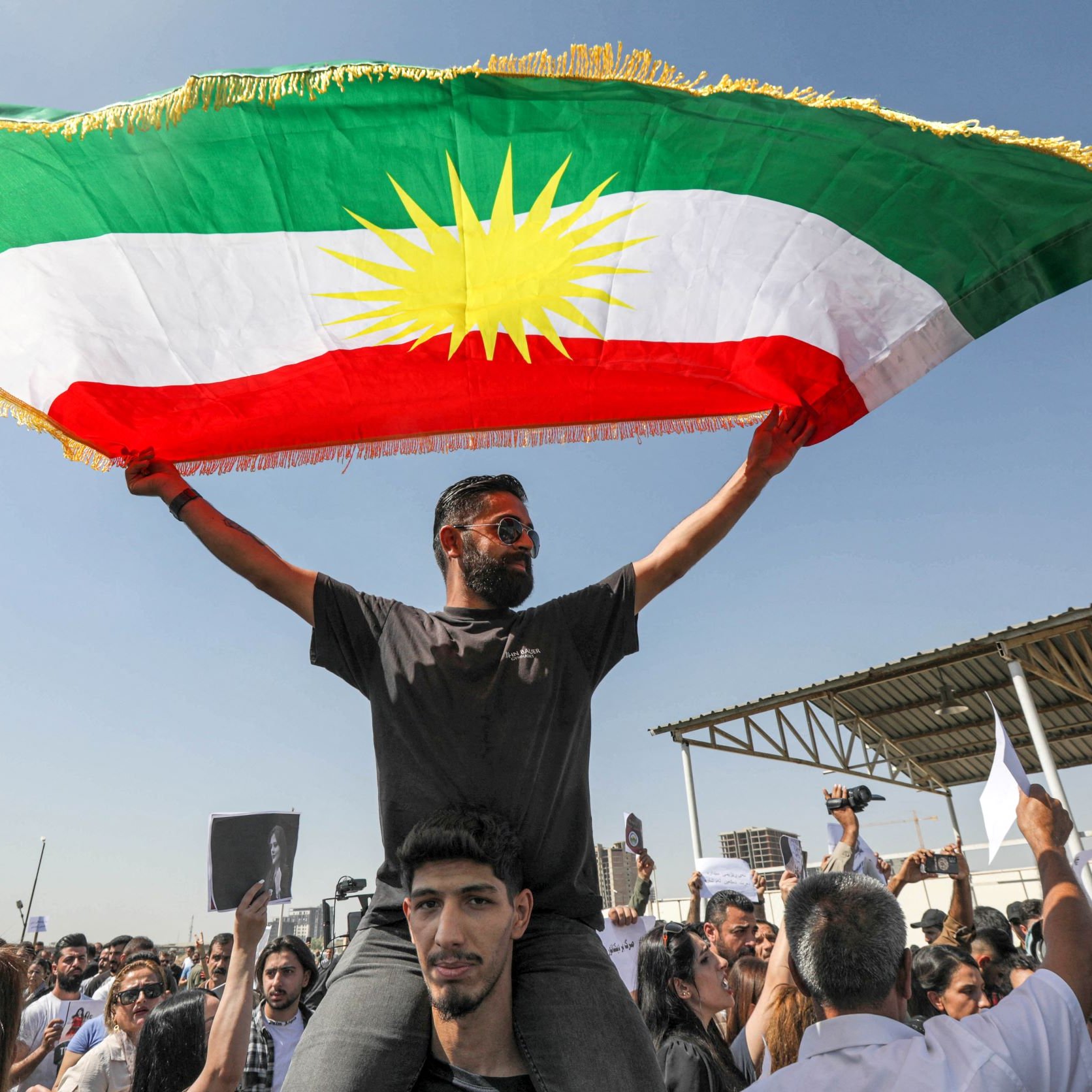 در این تصویر، فردی را مشاهده می‌کنیم که در بالای شانه‌های یک شخص دیگر ایستاده و پرچم کردستان را به اهتزاز درآورده است. پرچم دارای رنگ‌های سبز، سفید و قرمز با خورشید زرد در وسط است. جمعیتی در اطراف آنها حضور دارد و به نظر می‌رسد در حال برگزاری یک تظاهرات یا تجمع هستند. فضای تصویر آکنده از انرژی و احساسات جمعی است.