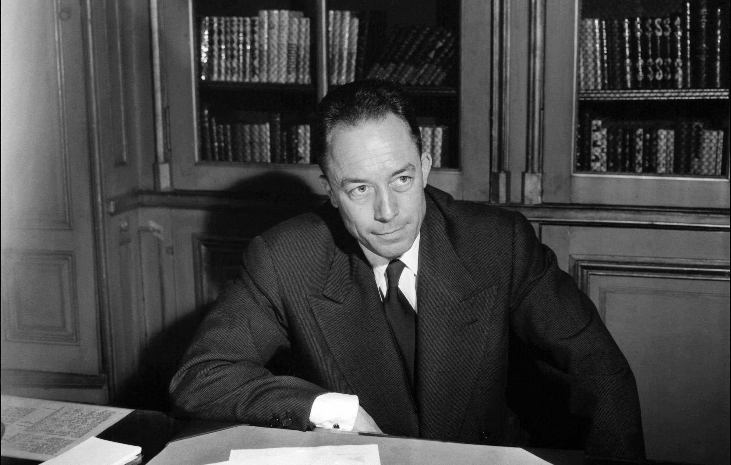 L'immagine mostra un uomo seduto a un tavolo, con dei documenti sparsi davanti a lui. Indossa un abito scuro con una cravatta, e appare pensieroso mentre guarda verso un punto oltre la fotocamera. Sullo sfondo si intravedono alcune librerie con libri. L'illuminazione è elegante, creando un'atmosfera seria e riflessiva.