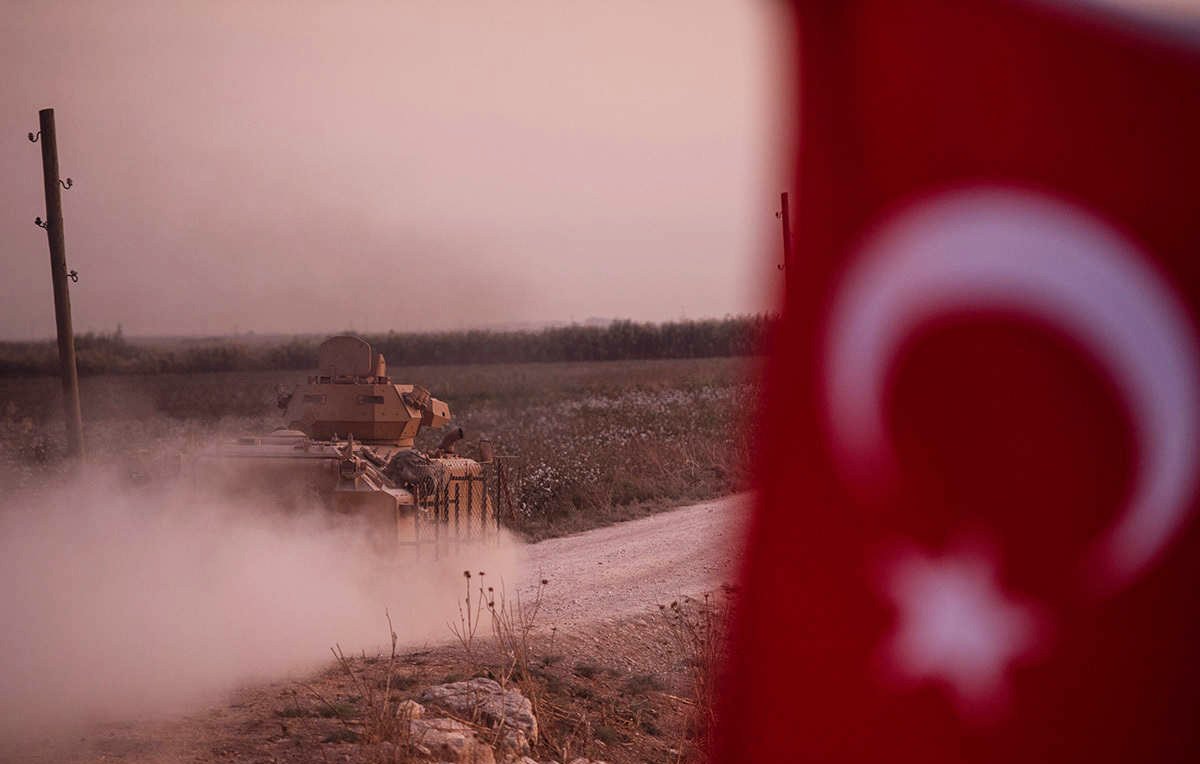در این تصویر، یک تانک نظامی در حال حرکت در یک جاده خاکی به سمت جلو دیده میشود. در پسزمینه، میدانهای سبز و طبیعتی آرام قرار دارد. در سمت چپ تصویر، پرچم ترکیه با نشانههای مشخص آن قرار دارد. غبار و دود ناشی از حرکت تانک فضای تصویر را پوشانده و حس تحرک و فعالیت نظامی را منتقل میکند. نور ملایم غروب یا صبح نیز به فضای کل تصویر جذابیت بیشتری بخشیده است.