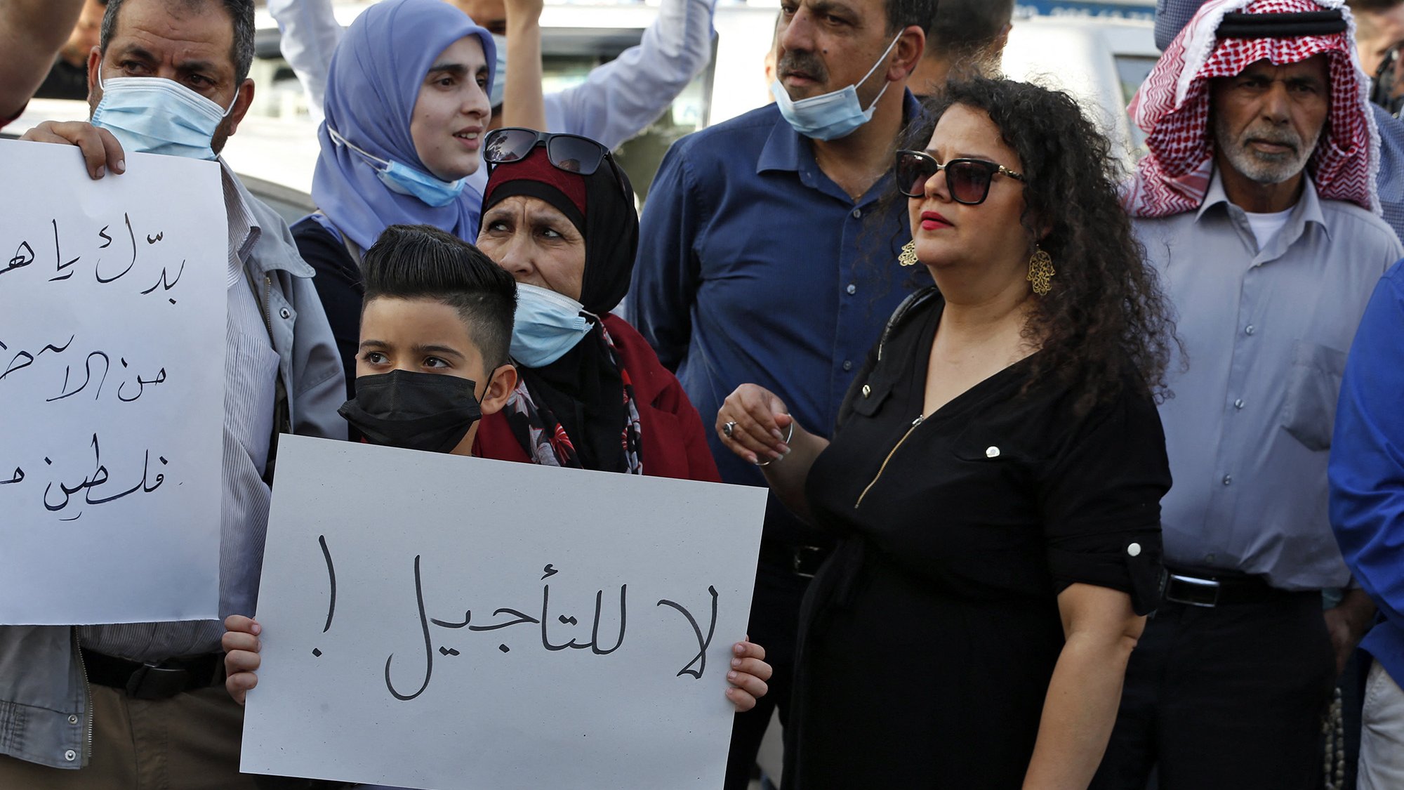 تظهر في الصورة مجموعة من الناس في مظاهرة. يرفع بعضهم لافتات، بينما يرتدي الآخرون كمامات. في المقدمة، يوجد طفل صغير يحمل لافتة مكتوب عليها "لا للتأجيل!"، وتحيط به مجموعة من البالغين، بينهم امرأة ترتدي فستانًا أسود وبعض الرجال بملابس تقليدية. تعكس الصورة جوًا من الاحتجاج والتعبير عن الرأي.
