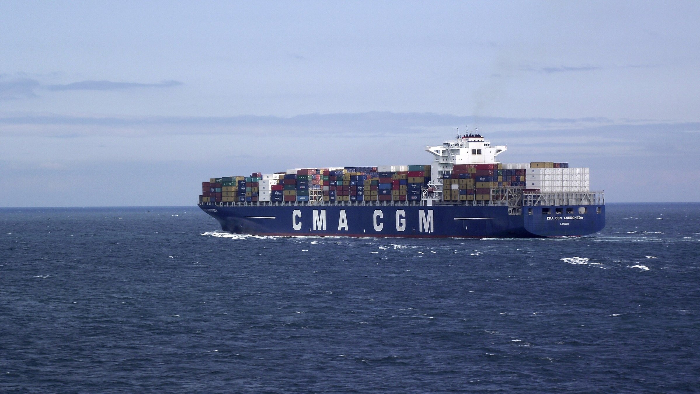 L'image montre un grand porte-conteneurs naviguant sur une mer calme. Le navire, qui porte les lettres "CMA CGM" sur son flanc, est couvert de conteneurs multicolores empilés sur le pont. Le ciel est légèrement nuageux, avec des teintes de bleu, et on peut apercevoir des vagues sur l'eau. L'ensemble dégage une impression de commerce maritime et de transport international.