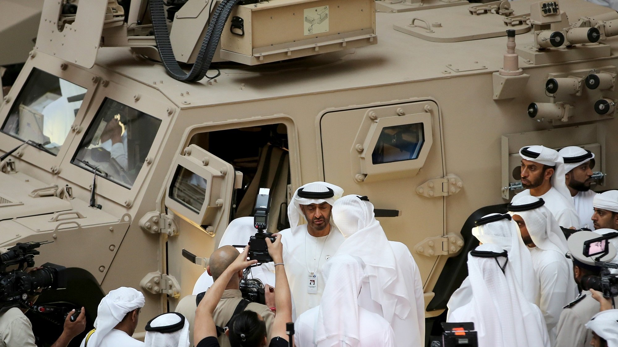 در این تصویر، گروهی از مردان به لباس سنتی عربی، در حال برگزاری یک رویداد یا نمایشگاه هستند. یکی از افراد در مرکز تصویر به نظر می‌رسد که در حال صحبت با دیگران است و چند نفر دیگر نیز در حال عکس‌برداری هستند. در پس‌زمینه، یک وسیله نقلیه نظامی با رنگ شن و طراحی خاص دیده می‌شود. این صحنه نشان‌دهنده یک موقعیت رسمی و احتمالا نظامی است، با حضور رسانه‌ها و توجه به جزئیات.