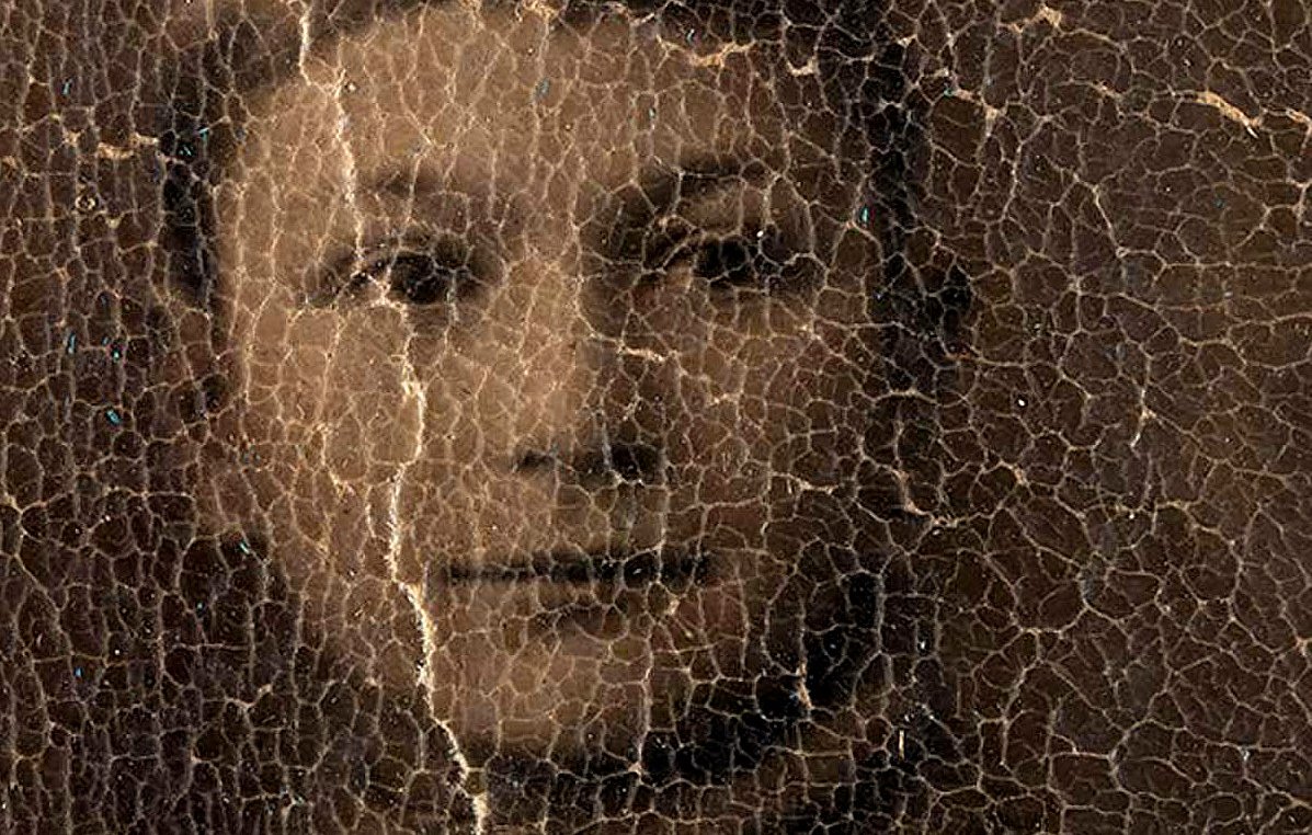 L'image représente un portrait d'une personne, probablement un jeune garçon, dont les traits sont légèrement obscurcis par un certain vieillissement et des fissures à la surface. Le visage montre des expressions délicates et le regard engageant, mais les détails sont atténués à cause de l'état usé de la photographie. Le fond semble sombre, ce qui accentue l'apparence vieillie de l'image.