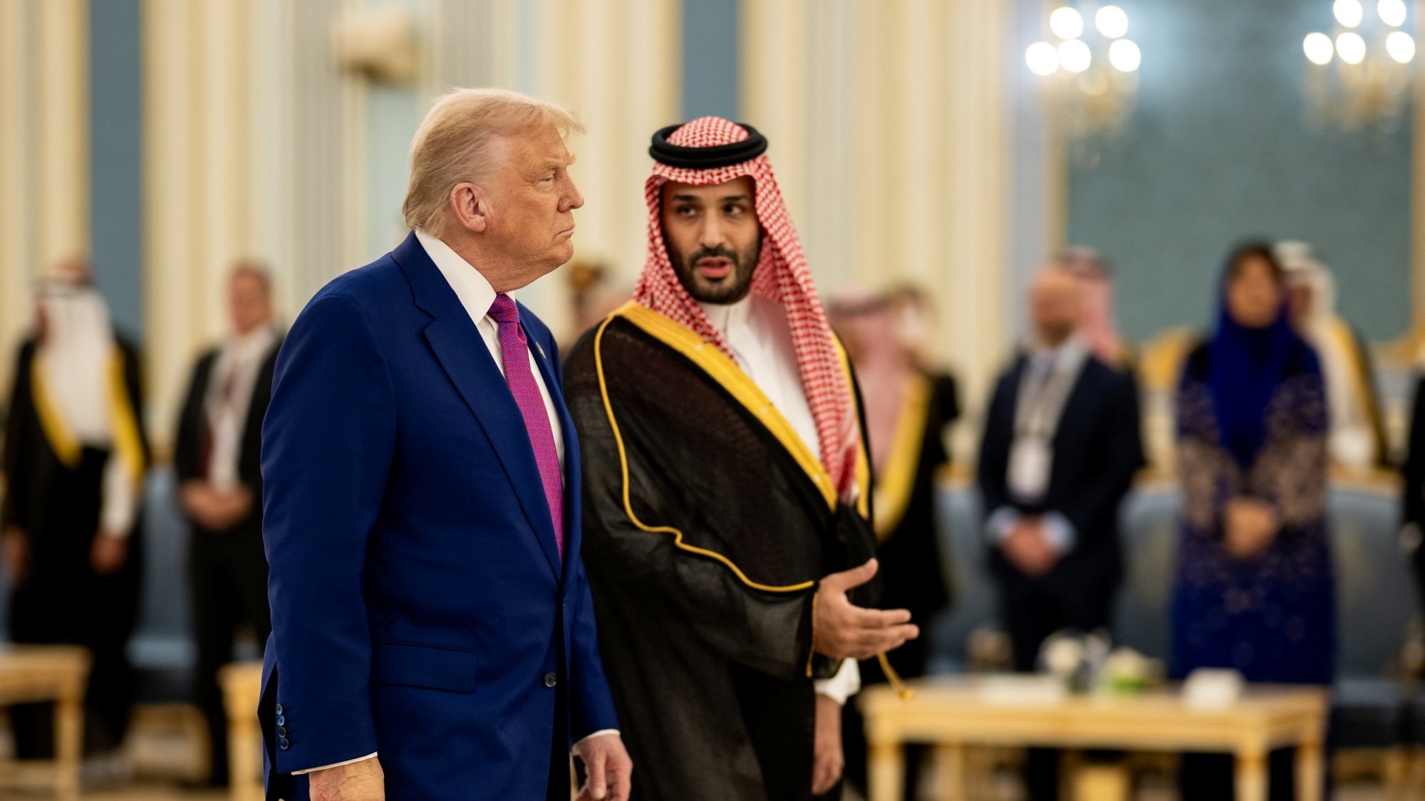 دو مرد در لباس‌های رسمی مختلف در یک محیط مجلل با نور ملایم و حضور افرادی دیگر.