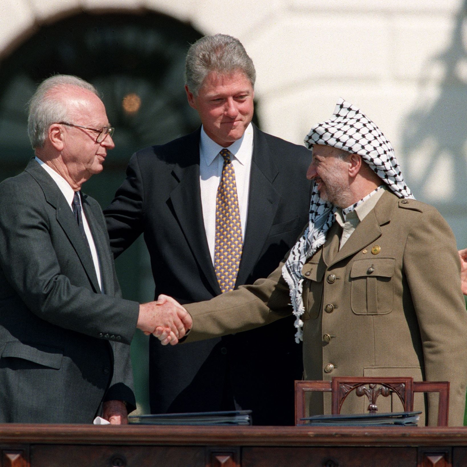 L'immagine mostra un momento storico di un incontro tra tre figure di spicco: un uomo con occhiali e capelli grigi che stringe la mano a un altro uomo vestito con un abito scuro, mentre un terzo uomo, vestito con un'uniforme militare e un copricapo tradizionale, osserva con un sorriso. La scena si svolge in un contesto ufficiale, probabilmente durante una cerimonia importante, e c'è un'atmosfera di amicizia e collaborazione. Sullo sfondo si possono scorgere elementi architettonici di un edificio governativo.