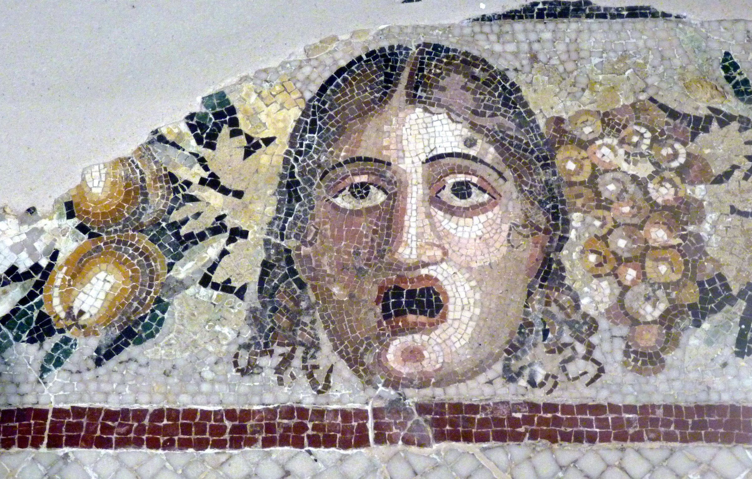 L'image présente un ancien mosaïque représentant un visage humain. Le caractère exprime une émotion marquée, semblant étonné ou effrayé. Autour du visage, il y a des éléments décoratifs, notamment des grappes de raisin et des fruits, qui ajoutent une touche colorée et contrastent avec le fond neutre de la mosaïque. La technique utilisée évoque la richesse de l'art ancien et son attention aux détails.