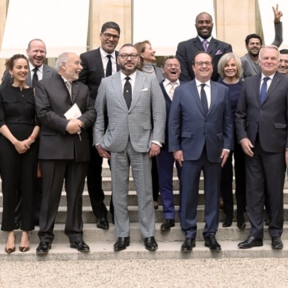 L'image montre un groupe de personnes posant sur des marches devant un bâtiment, probablement lors d'un événement officiel. Au centre, deux hommes se tiennent côte à côte, avec des costumes élégants. Les autres membres du groupe sont habillés de manière formelle ou semi-formelle et sourient pour la photo. L'ambiance semble conviviale, et l'un des hommes à l'arrière fait un signe de paix avec sa main. Les lieux sont lumineux et semblent bien entretenus, ajoutant à l'atmosphère de l'événement.