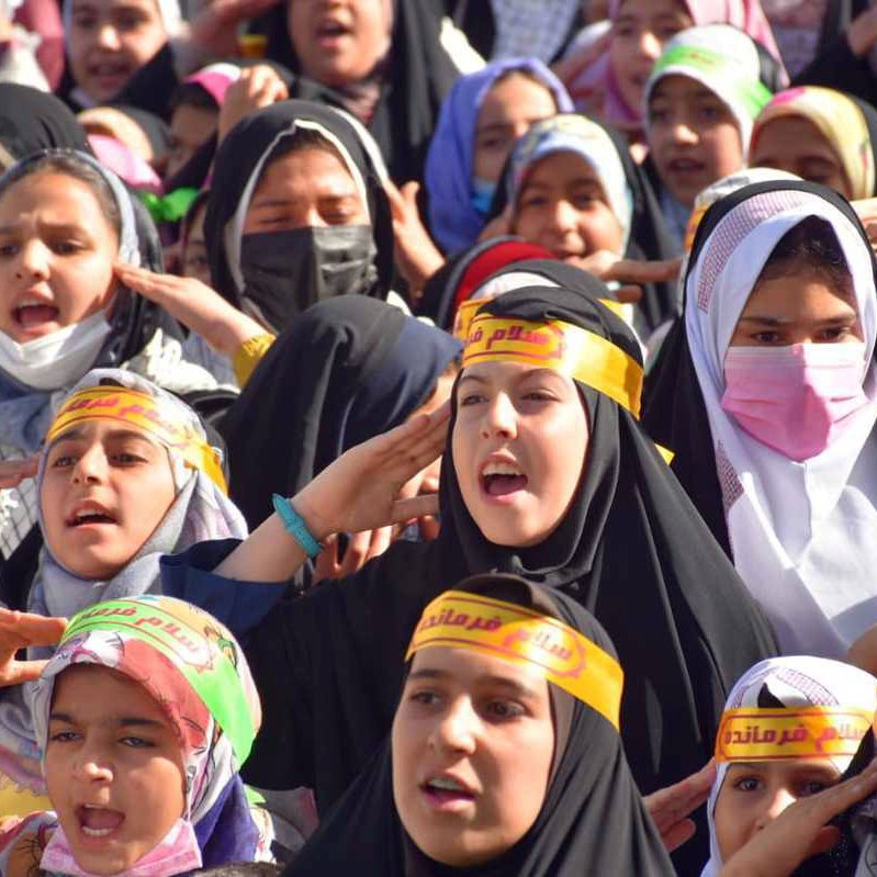 The image shows a large group of girls and young women gathered together, likely participating in an event or demonstration. They are wearing colorful headscarves and black clothing, along with yellow headbands that feature Arabic text. The girls appear to be actively engaged, singing or chanting, and expressing enthusiasm. Their expressions convey a sense of unity and purpose as they participate in this collective activity.
