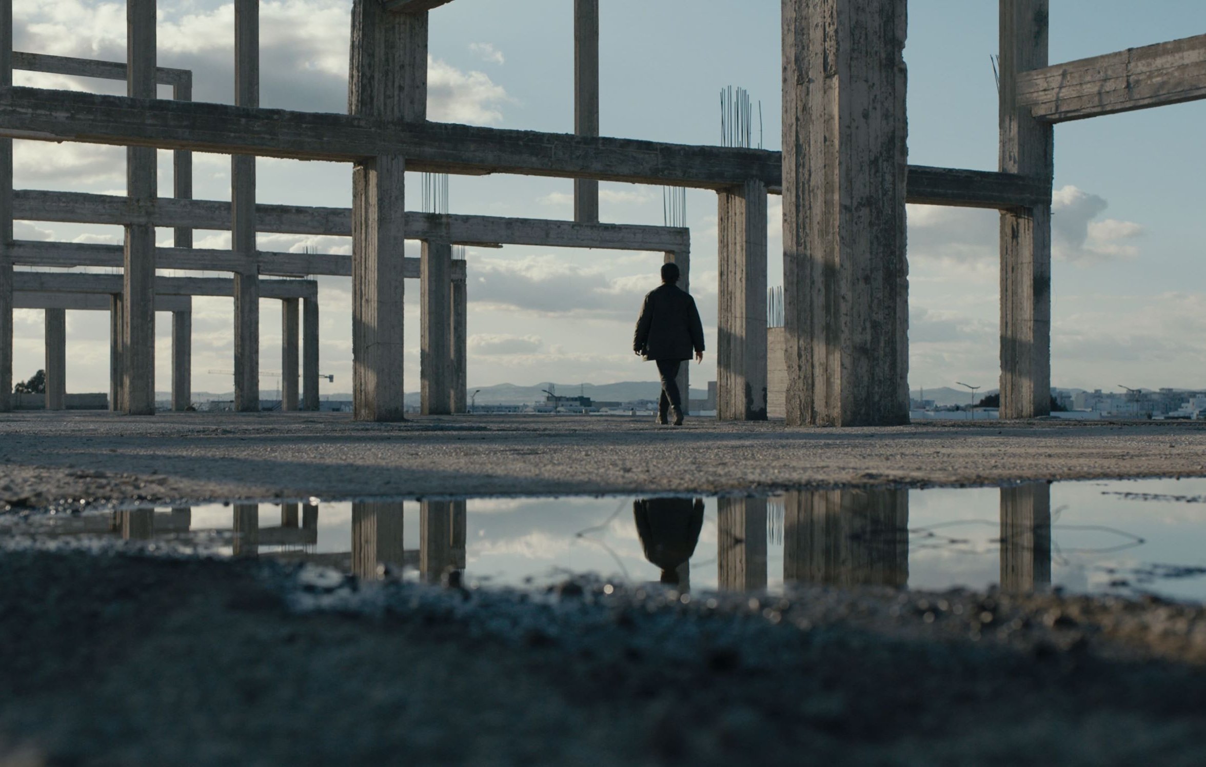 L'image montre une personne marchant à travers une structure de bâtiment inachevée. Les piliers en béton se dressent autour d'elle, créant une atmosphère à la fois vide et mystérieuse. Le sol est humide, et une flaque d'eau reflète les piliers et le ciel, ajoutant une dimension visuelle intéressante à la scène. Le ciel est partiellement nuageux, et l'éclairage est doux, contribuant à l'ambiance contemplative de cet espace abandonné.