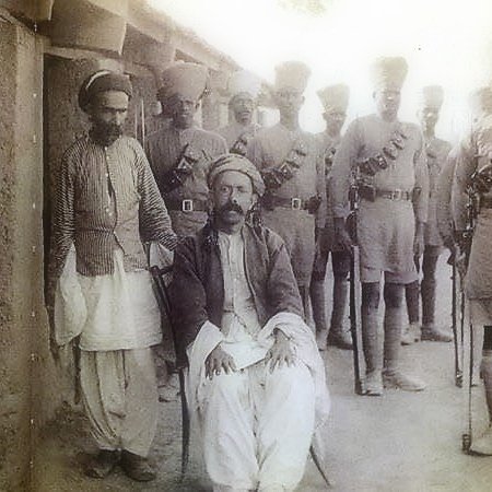 L'image montre un groupe de personnes en tenue traditionnelle, avec un homme assis au premier plan, semble-t-il en position plus détendue. Il porte des vêtements amples et un turban. Autour de lui, plusieurs hommes en uniforme militaire se tiennent debout, équipés de ceintures et de fusils, donnant une impression de formation ou de cérémonie. L'environnement semble être un lieu extérieur, probablement historique.