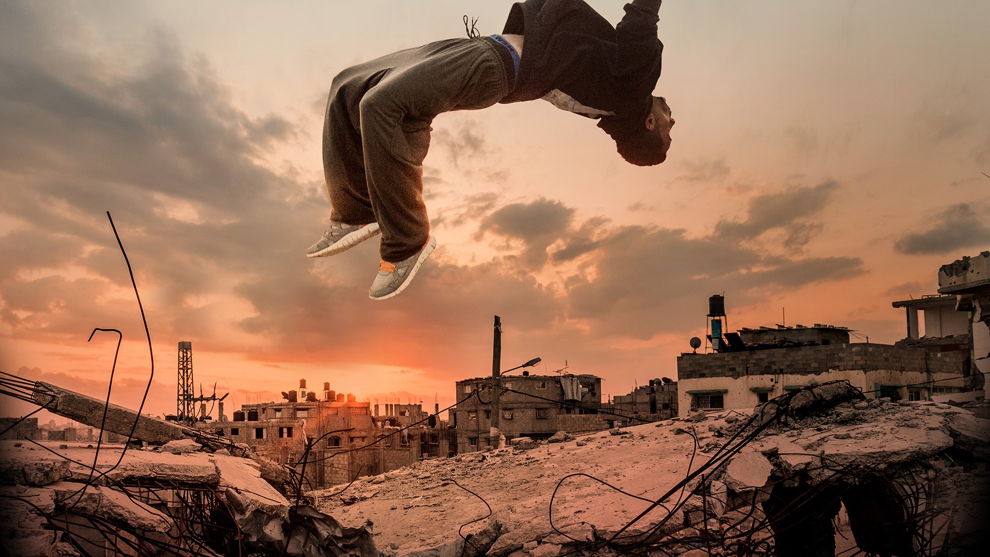 L'image montre un jeune homme en train de sauter dans les airs, effectuant une figure acrobatique, au-dessus des ruines d'une ville. En arrière-plan, le ciel est coloré de teintes orangées et bleutées, ce qui donne une ambiance dramatique à la scène. Les débris et les structures en ruine témoignent d'un environnement urbain dégradé, ajoutant un contraste frappant avec la dynamique du saut.