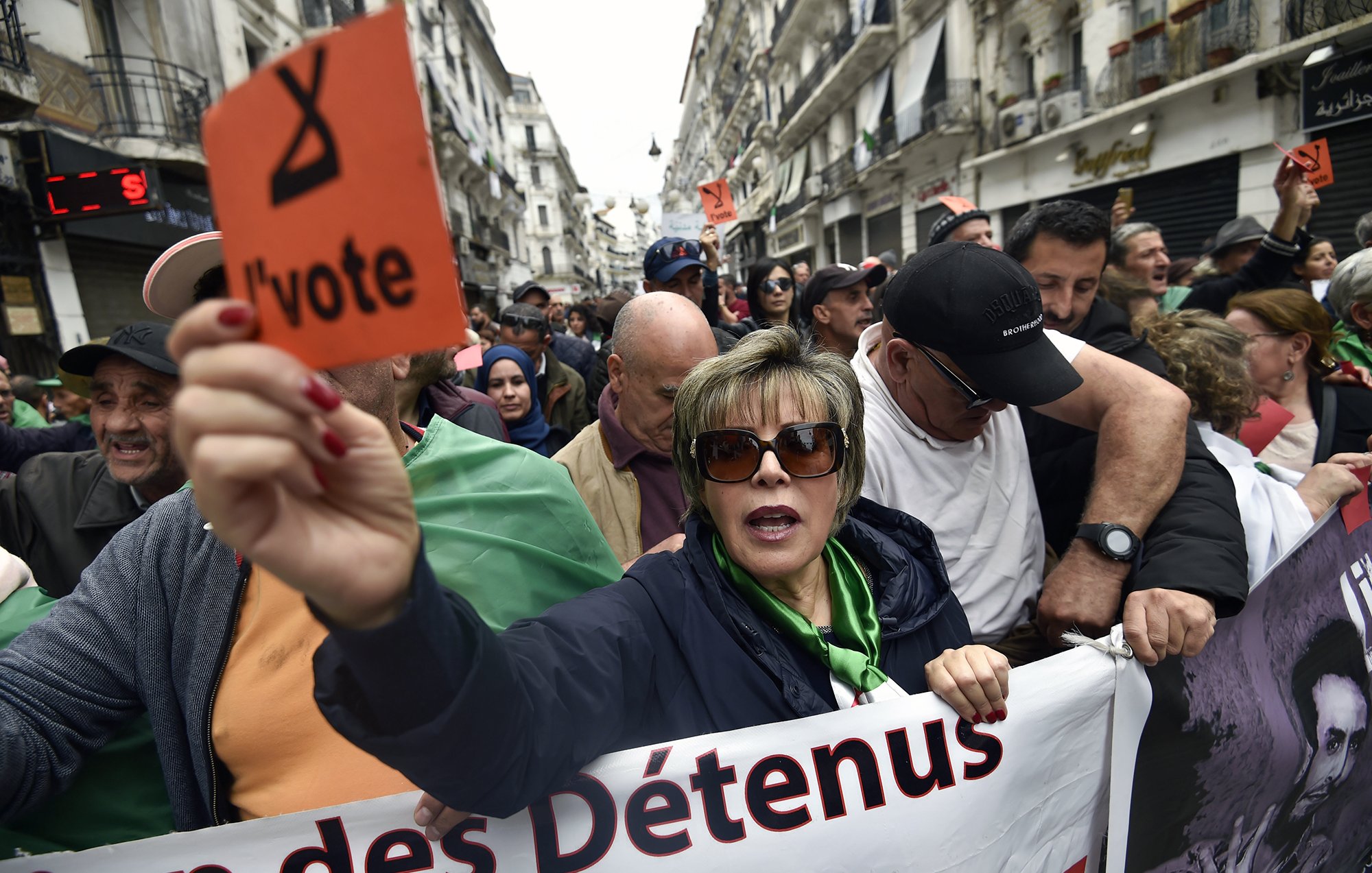 L'image montre une manifestation dans une rue d'Alger. Au premier plan, une femme tient une pancarte orange avec le mot "l'vote". Elle est entourée par d'autres manifestants, dont certains portent des drapeaux algériens. L'atmosphère semble engagée et déterminée, avec des expressions de solidarité parmi les participants. Des banderoles sont visibles, indiquant des revendications liées à la situation politique. L'environnement urbain et les bâtiments en arrière-plan témoignent d'une ambiance dynamique de protestation.