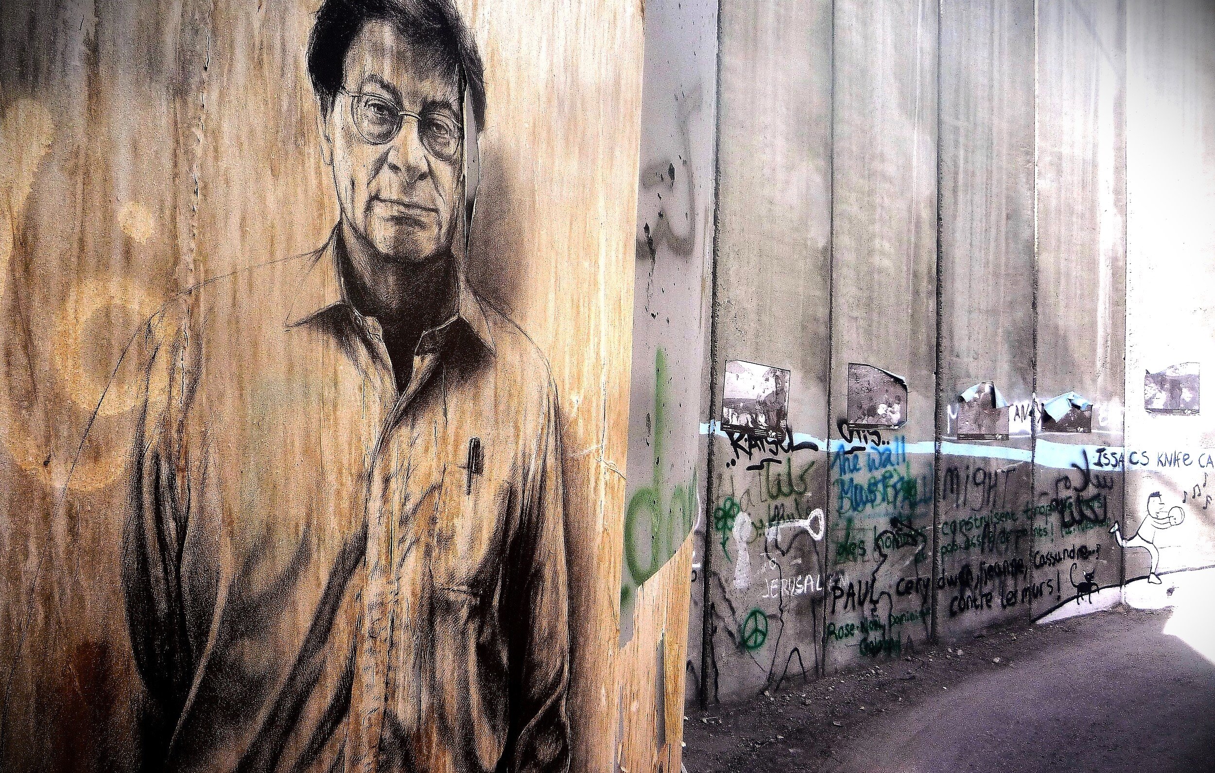 L'immagine rappresenta un murale su un muro di cemento, caratterizzato da un ritratto di un uomo con occhiali e un'espressione seria. Il muro è grezzo e presenta anche scritte e graffiti, suggerendo un contesto urbano e di protesta. Il contrasto tra il ritratto e il resto del muro evidenzia un messaggio artistico e sociale. La scena evoca un senso di riflessione e di critica.