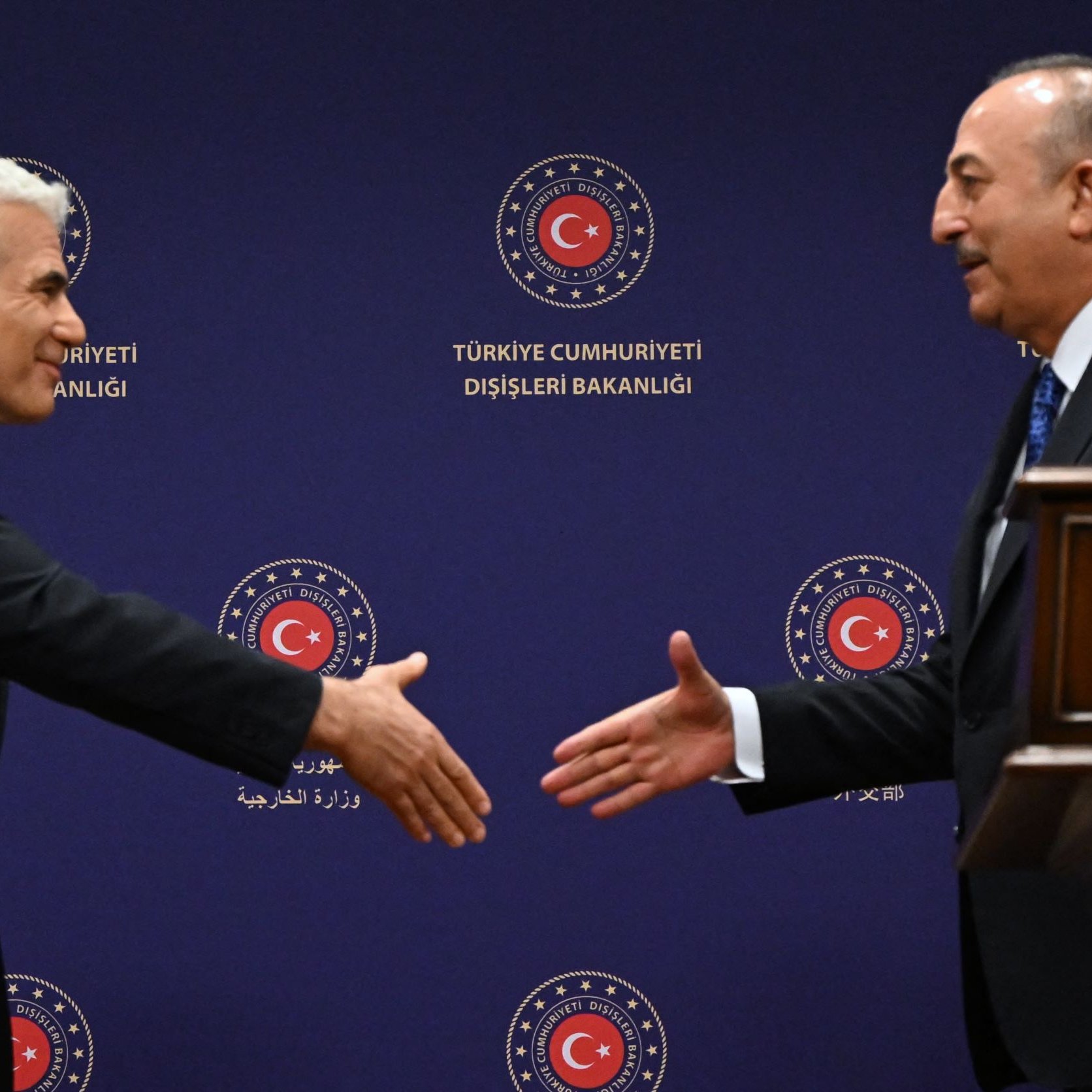 L'image montre deux hommes se préparant à se serrer la main dans un cadre officiel. Ils se tiennent devant un fond avec des symboles de la République de Turquie et des inscriptions officielles. L'un des hommes porte un costume sombre et semble sourire, tandis que l'autre a également un costume et une expression sérieuse. L'atmosphère dégage une impression de diplomatie et de protocole.