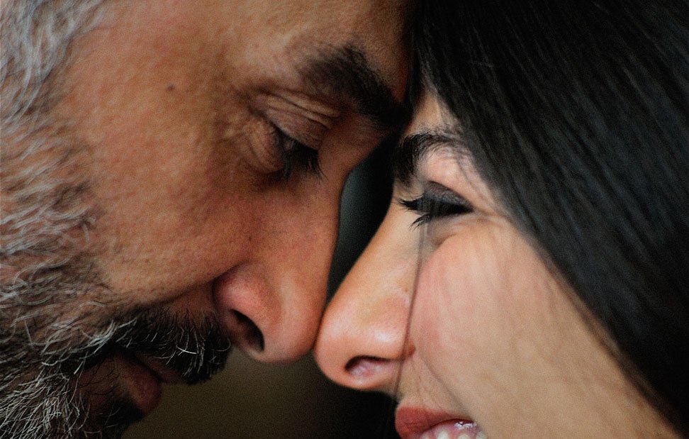 Cette image montre deux personnes, un homme et une femme, qui sont très proches l'un de l'autre. Ils touchent leurs fronts et semblent partager un moment intime. L'homme a une barbe grisonnante, tandis que la femme a des cheveux longs et foncés. Leur expression est douce : l'homme a un air serein, et la femme sourit joyeusement. L'ambiance de l'image évoque l'affection et la complicité entre les deux individus.
