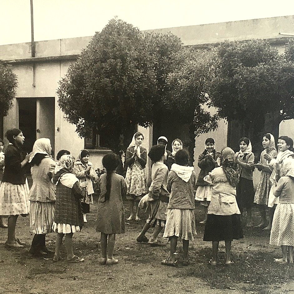 تظهر في الصورة مجموعة من الفتيات والأطفال يلعبون في ساحة مفتوحة. يبدو أنهن يرتدين ملابس تقليدية، وبعضهن يحمل غطاء رأس. الفتيات محاطة بأشجار، مما يضفي جوًا طبيعيًا على المكان. يبدون مشغولات باللعب والضحك، في مشهد يعكس روح الطفولة والنشاط. الخلفية بسيطة، مع وجود مبنى مكون من جدران بسيطة.