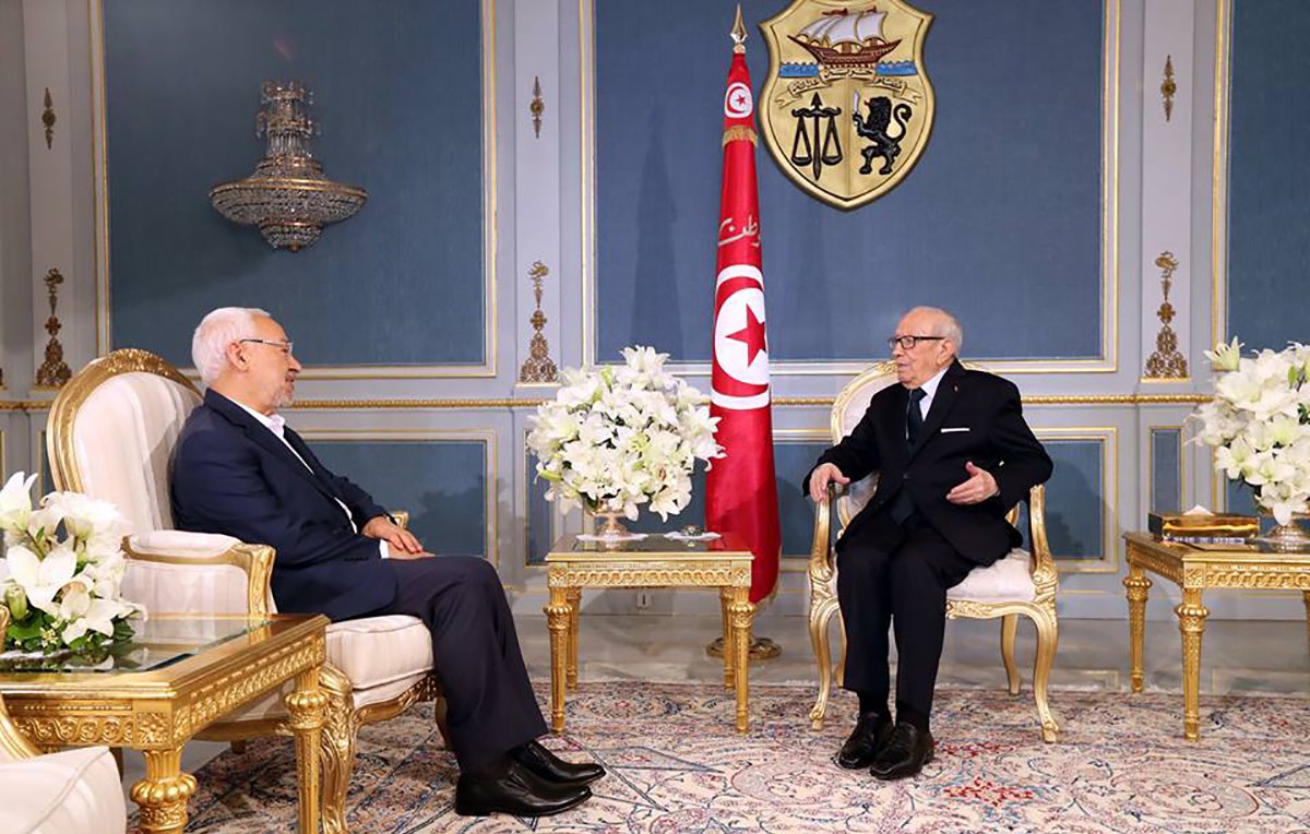 L'image montre deux hommes assis dans un cadre officiel, probablement dans un bureau ou une salle de réunion élégante. Les deux sont vêtus de costumes sombres, l'un portant des lunettes. Au fond, on peut voir le drapeau tunisien ainsi qu'un blason. La pièce est décorée avec des meubles en or, des fleurs blanches et un tapis coloré, ce qui crée une atmosphère formelle et importante.