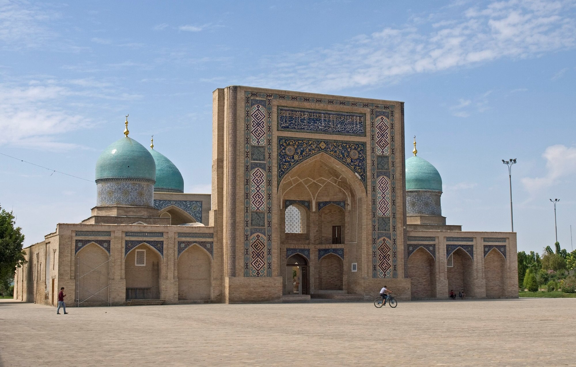 Bâtiment en brique avec de grandes arches et coupoles turquoise sous un ciel bleu.