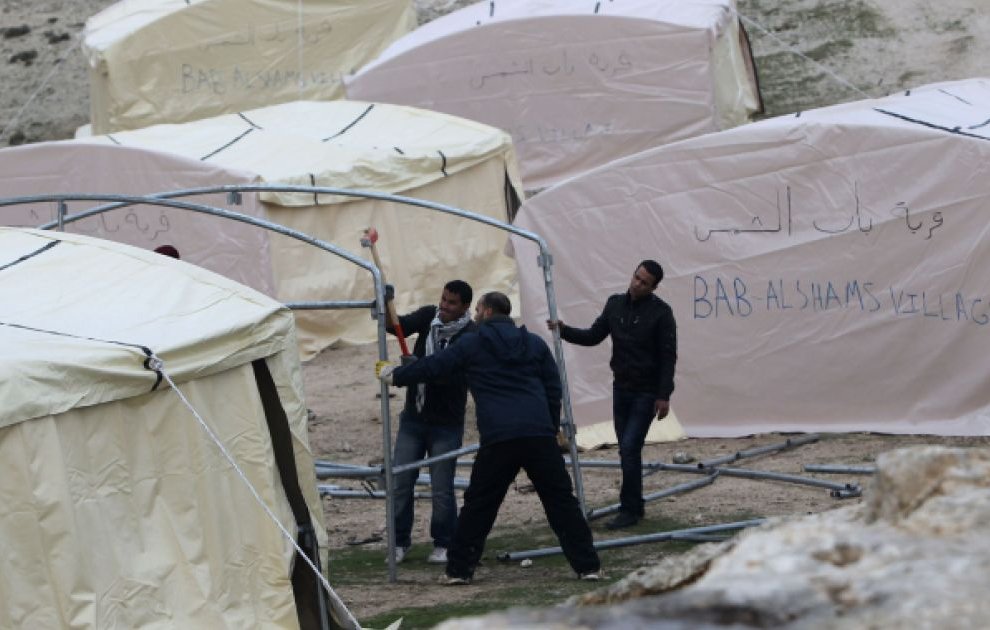 L'image montre plusieurs tentes de type campement, qui semblent être installées dans un environnement extérieur. Les tentes ont des écrits en arabe et en anglais, mentionnant "BAB AL SHAMS VILLAGE". On observe également des individus en train de travailler ensemble pour monter les structures des tentes. L'ensemble de la scène évoque des activités de construction ou d'aménagement dans un cadre probablement temporaire.