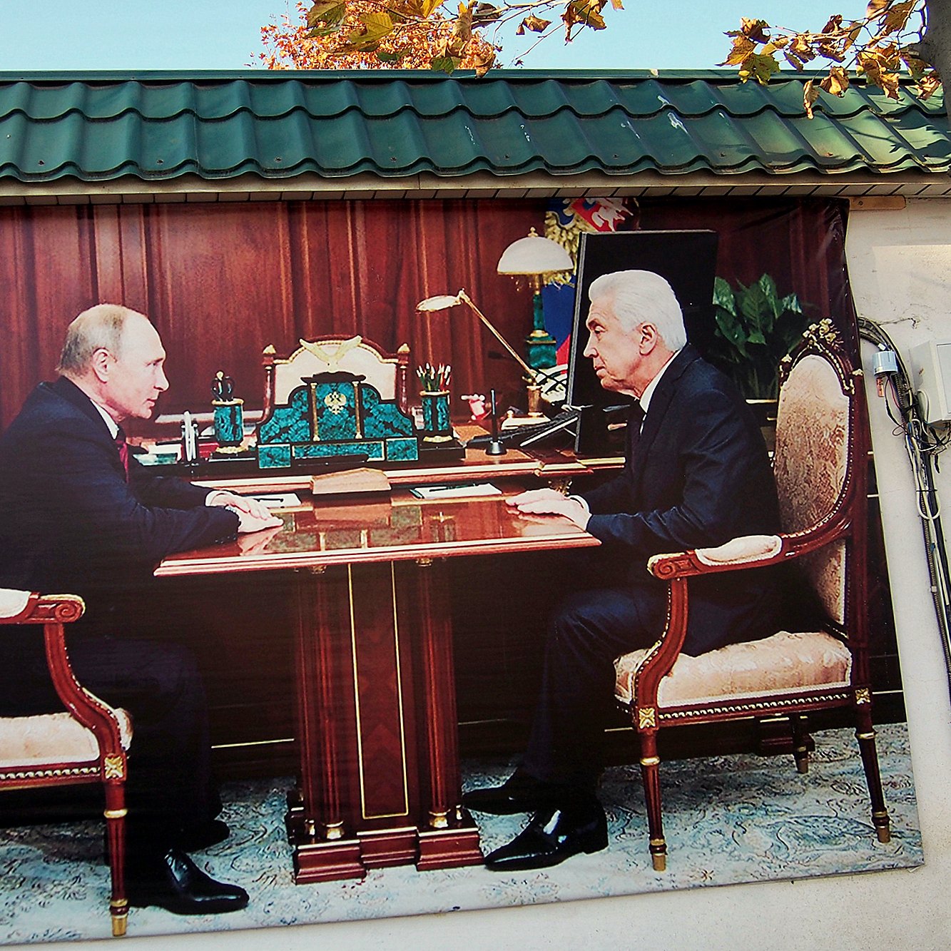 تظهر الصورة مشهدًا لمكتب يتواجد فيه شخصان يجلسان على جانبي طاولة كبيرة، يبدو أنهما يتحدثان أو يتناقشان. الخلفية تحتوي على أثاث مكتبي فاخر، مع تفاصيل مثل المصابيح والأدوات المكتبية. المنظر العام يعطي انطباعًا بالجدية والنقاش المهم بين الشخصين.