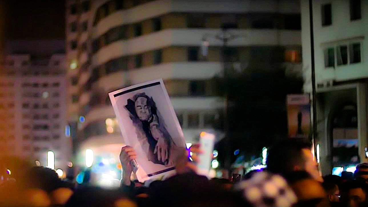 L'image montre une foule rassemblée lors d'une manifestation nocturne. Parmi les participants, une personne tient une grande affiche illustrant un portrait. Le fond de la scène est entouré de bâtiments éclairés, contribuant à l'atmosphère vibrante de l'événement. La photo semble capturer un moment important d'expression collective et de solidarité.
