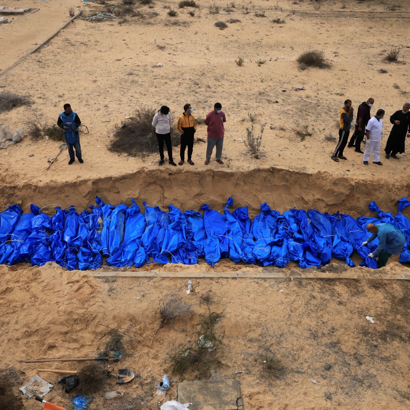 Des corps enveloppés de bleu sont placés dans une fosse, entourés de personnes en deuil.