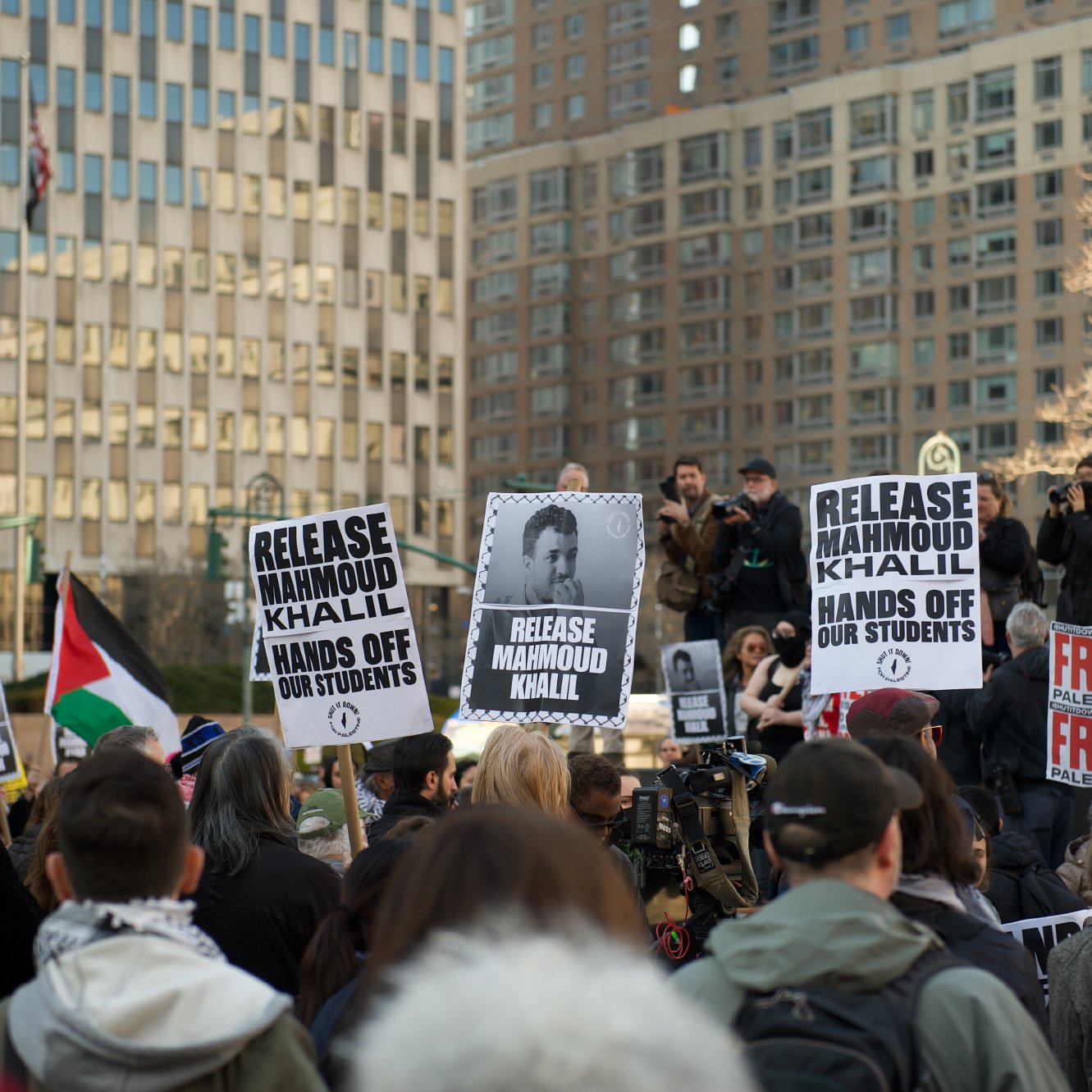 Des manifestants se rassemblent dans une ville, tenant des panneaux avec des slogans, notamment pour la libération de Mahmoud Khalil et des revendications pour les droits des étudiants. En arrière-plan, on peut voir des bâtiments et des personnes utilisant des caméras. L'ambiance est déterminée et engagée.