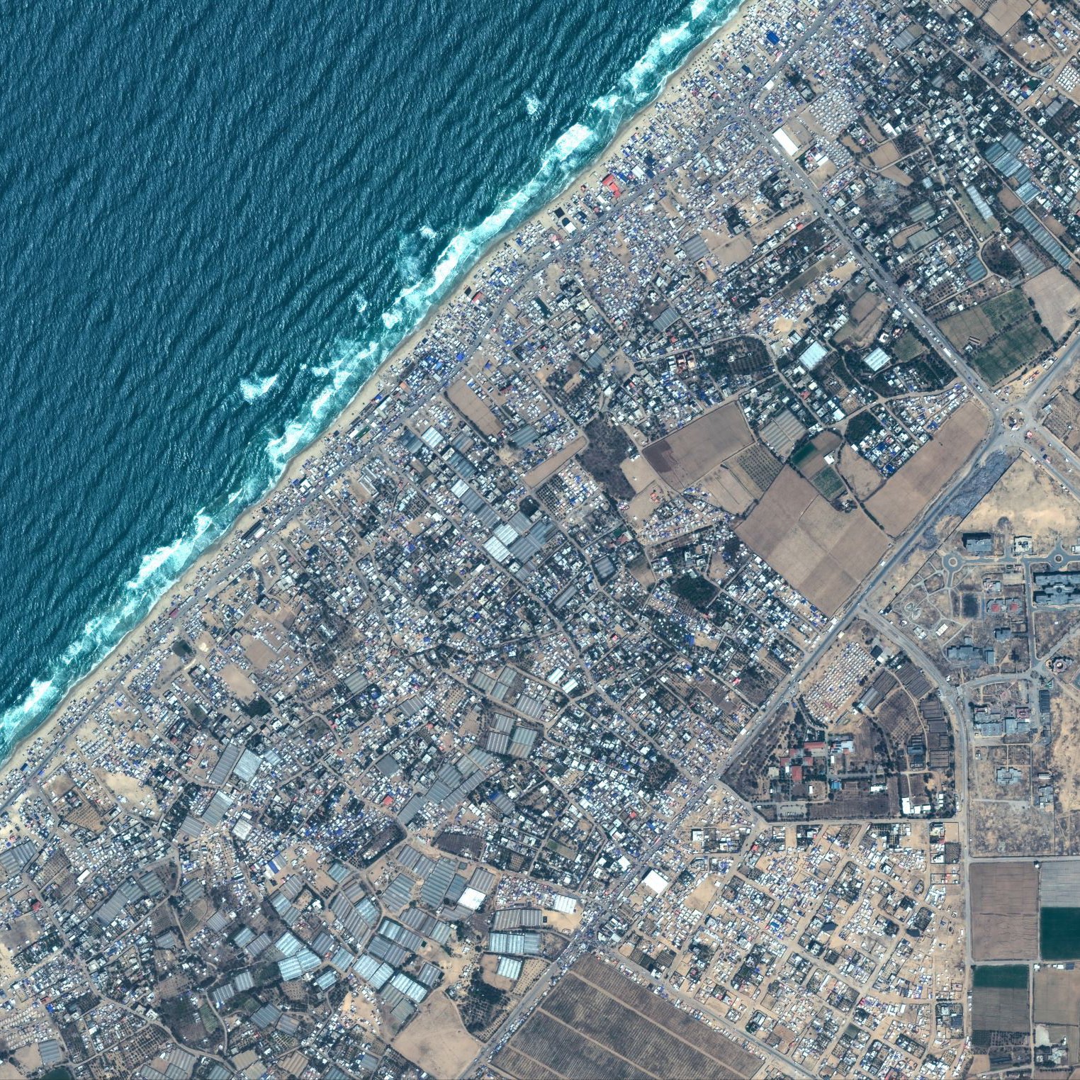 L'image montre une vue aérienne d'une zone côtière. En haut à gauche, on peut voir l'océan avec des vagues qui se brisent sur la plage. La partie inférieure de l'image présente des terrains variés, incluant des espaces urbanisés avec des routes et des bâtiments, ainsi que des zones agricoles avec des champs. La transition entre la zone bâtie et la nature est également visible. C'est une représentation d'un paysage où les zones urbaines rencontrent la côte.