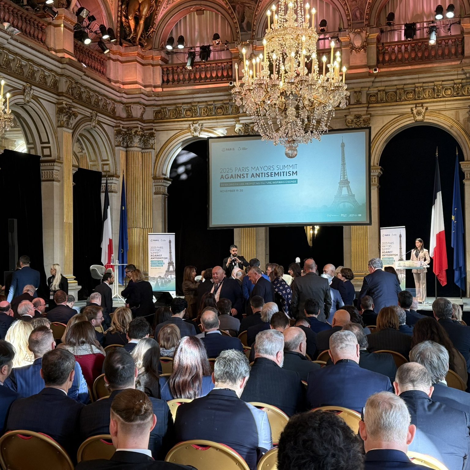 Grand hall avec chandeliers, foule réunie autour d'un discours contre l'antisémitisme.
