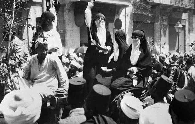 L'image montre une scène historique en noir et blanc, probablement dans une ville animée. On y voit plusieurs personnes, dont deux femmes en vêtements traditionnels ou religieux, tenant des positions centrales. Elles sont entourées d'une foule nombreuse qui semble attentive et engageante. L'atmosphère dégage un sentiment de mouvement et d'interaction, peut-être lors d'un événement culturel ou d'une célébration. Les traits vestimentaires et l'architecture en arrière-plan suggèrent une époque révolue.