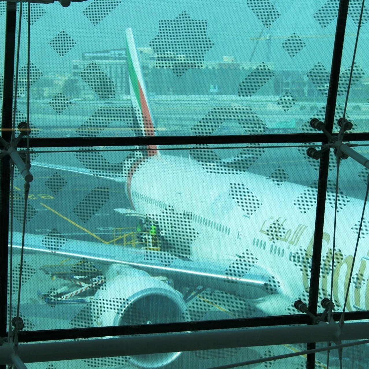 L'image montre un aéroport avec une vue à travers une grande fenêtre en verre. À l'extérieur, on peut apercevoir un avion de la compagnie Emirates, probablement un Boeing 777, stationné sur le tarmac. On distingue des travailleurs qui s'affairent autour de l'appareil, préparant son embarquement. La lumière ambiante semble assez diffuse, et il y a un léger flou qui donne une impression de profondeur. En arrière-plan, on peut voir d'autres infrastructures aéroportuaires.