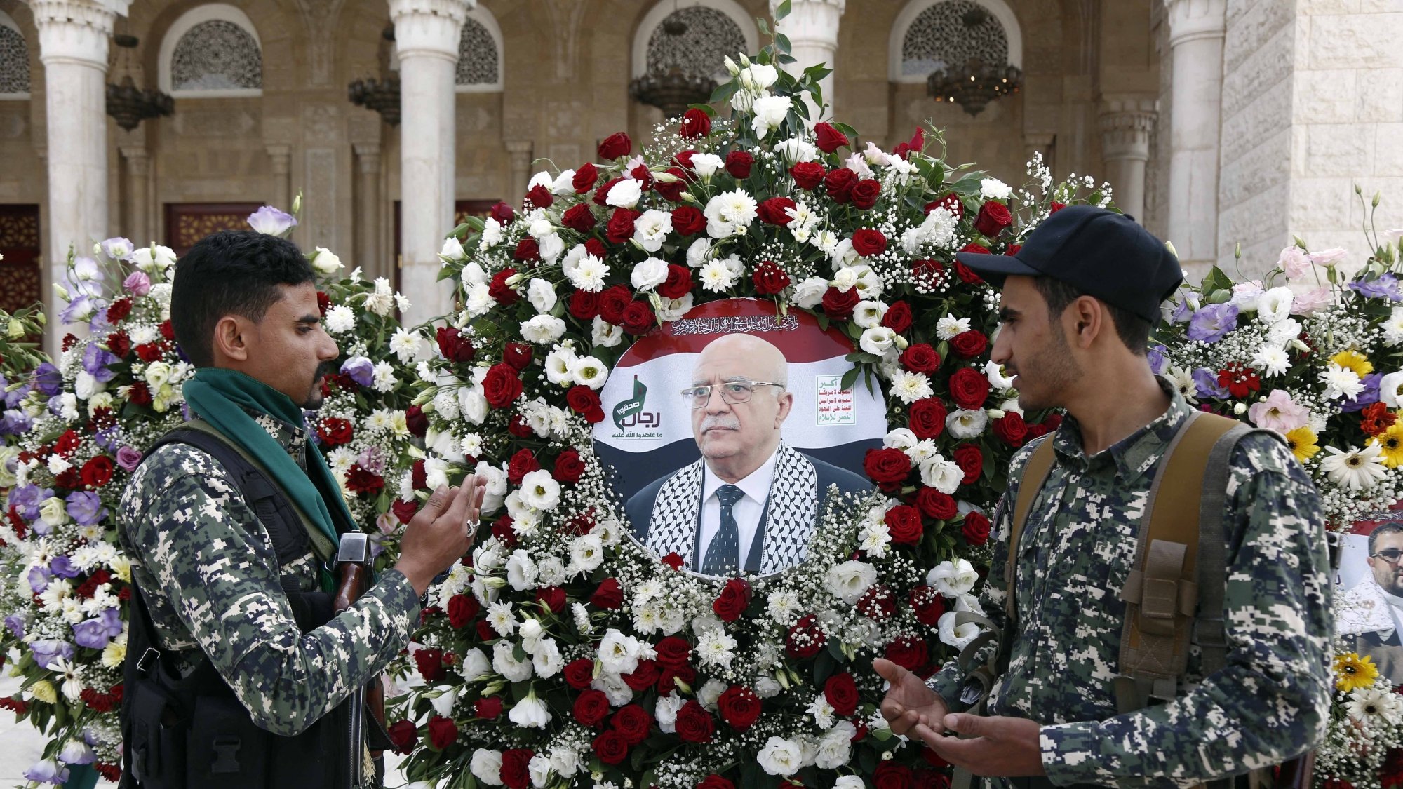 دو نظامی در کنار تاج گل با گل‌های قرمز و سفید ایستاده‌اند. تصویری از یک مرد وسط تاج قرار دارد.