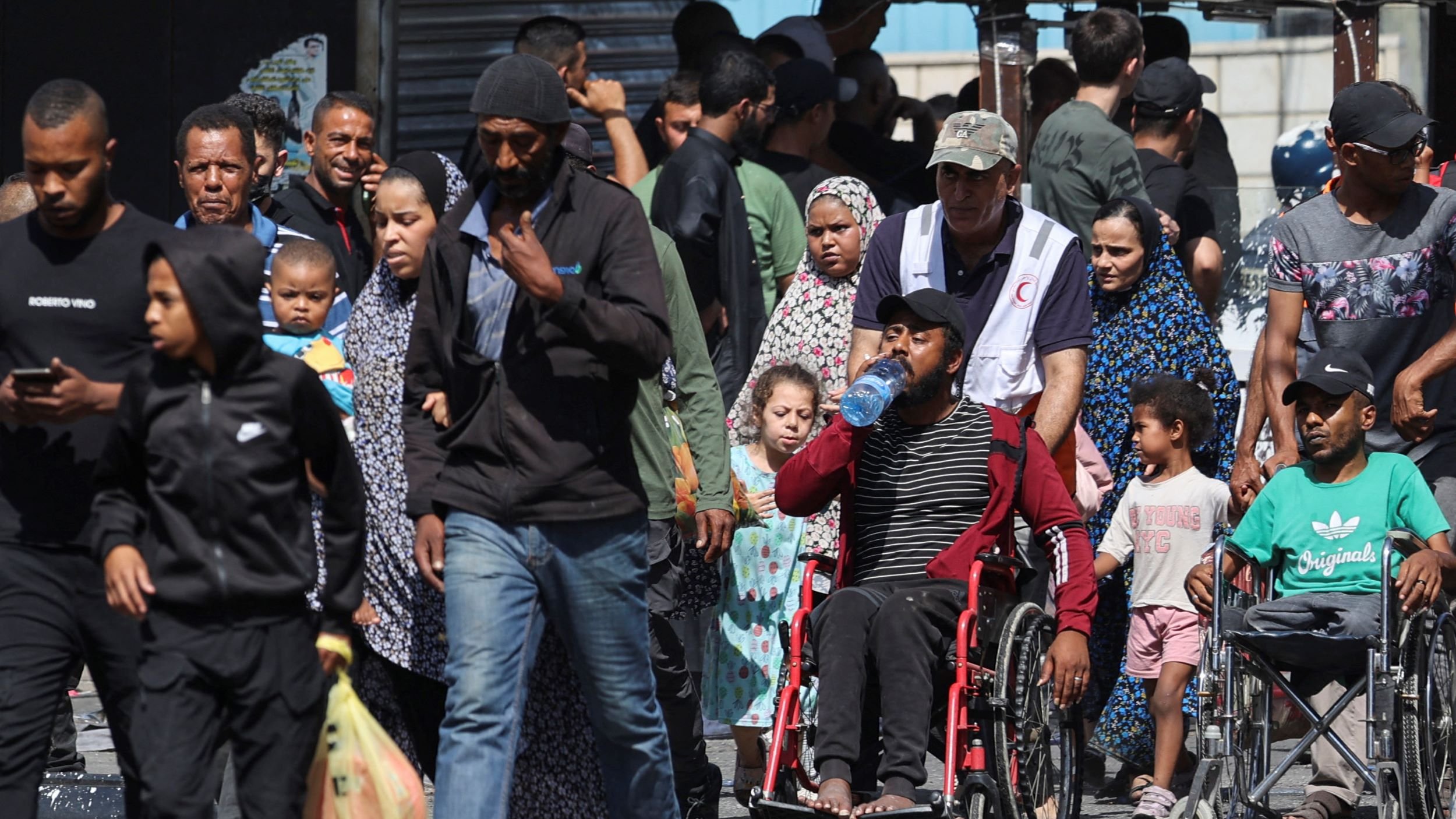 در این تصویر، گروهی از مردم در حال حرکت در یک خیابان شلوغ دیده میشوند. برخی از افراد در حال حمل کیف یا وسایل هستند و تعدادی از کودکان نیز در کنار بزرگترها دیده میشوند. فردی که روی ویلچر نشسته است به وضوح در مرکز تصویر قرار دارد و یک بطری آب در دست دارد. لباسهای افراد متنوع است و به نظر میرسد که برخی از آنها به طور مشخص به یک مراسم یا تجمع مربوط هستند. فضای تصویر از شلوغی و تنوع انسانها پر شده است.